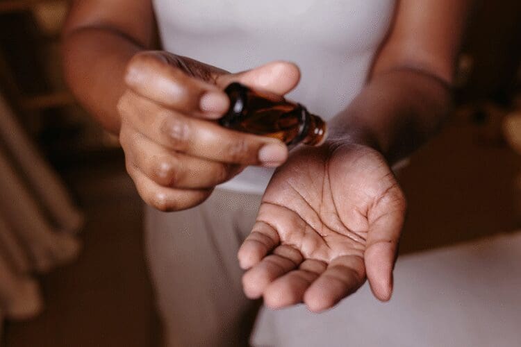 Person applying a cosmetic facial oil or serum from an amber glass bottle into the palm of their hand.