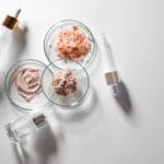 Cosmetic formulation ingredients with serum bottles, cream, minerals, and powder displayed in laboratory glass dishes.