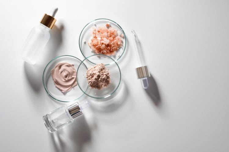 Cosmetic formulation ingredients with serum bottles, cream, minerals, and powder displayed in laboratory glass dishes.