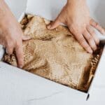 Person packing a cardboard box with recyclable paper filling, representing sustainable packaging and packaging compliance.