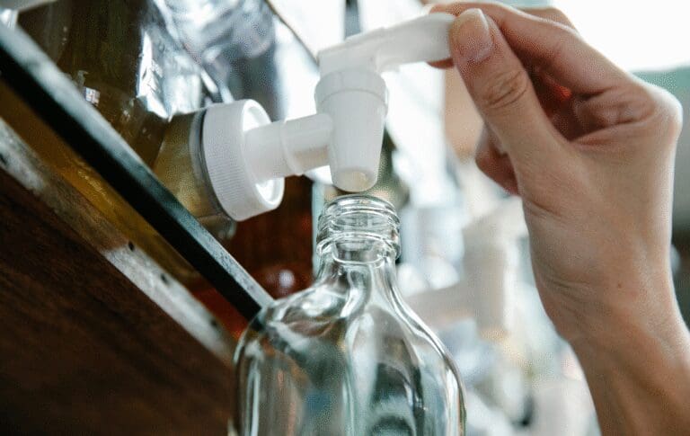 Person refilling a glass cosmetic bottle from a dispenser, representing reusable packaging and sustainable cosmetics.